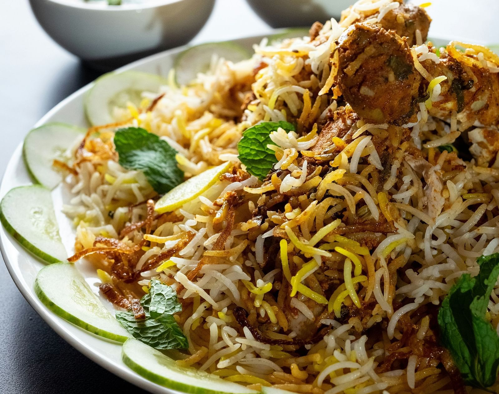 Aromatic chicken biryani served on a white plate, garnished with mint leaves, lemon slices, and cucumber, accompanied by raita and curry bowls.