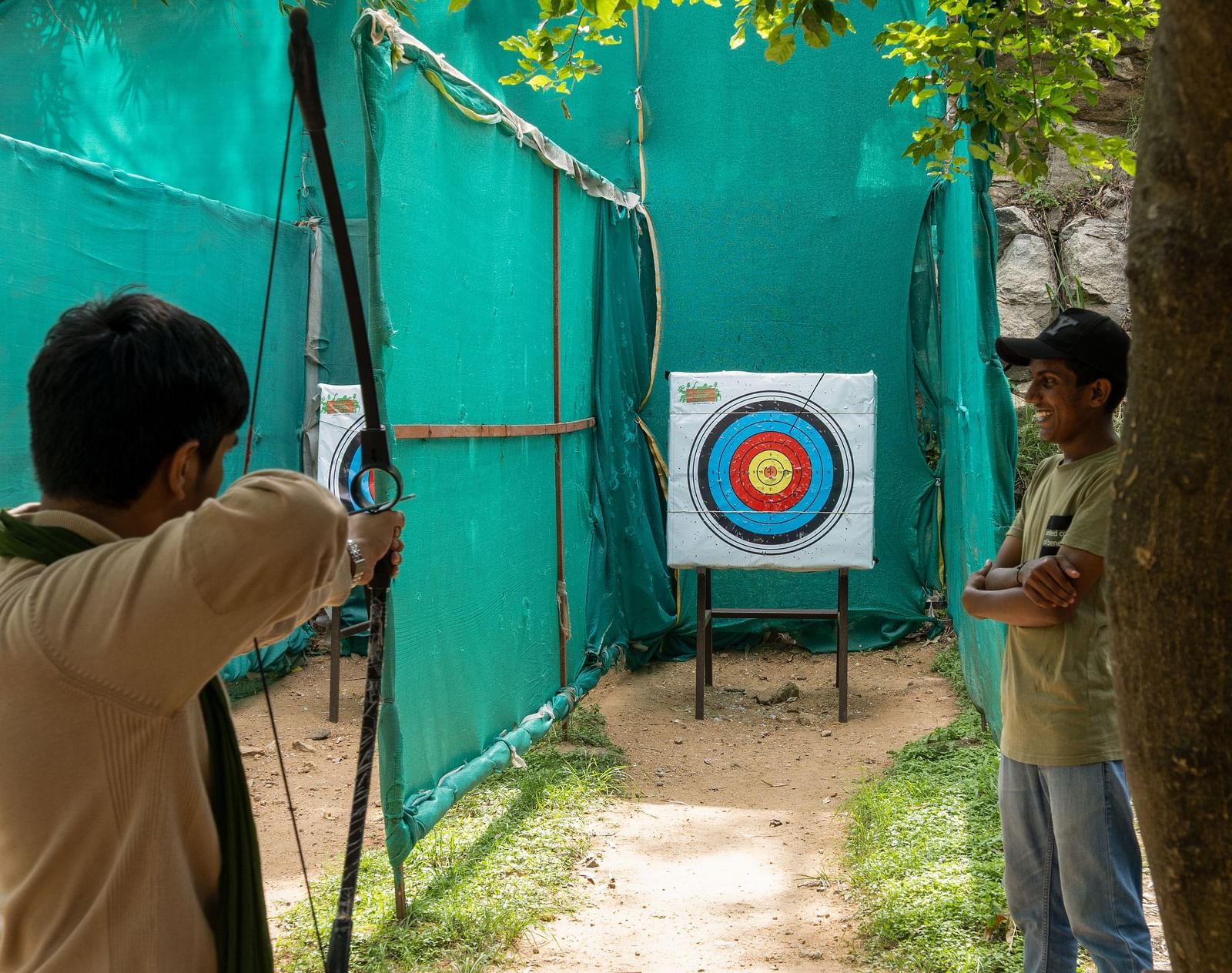 A person aims a bow and arrow at a target in an outdoor archery range, partially enclosed by green netting | RD's Nature Retreat, Bangalore