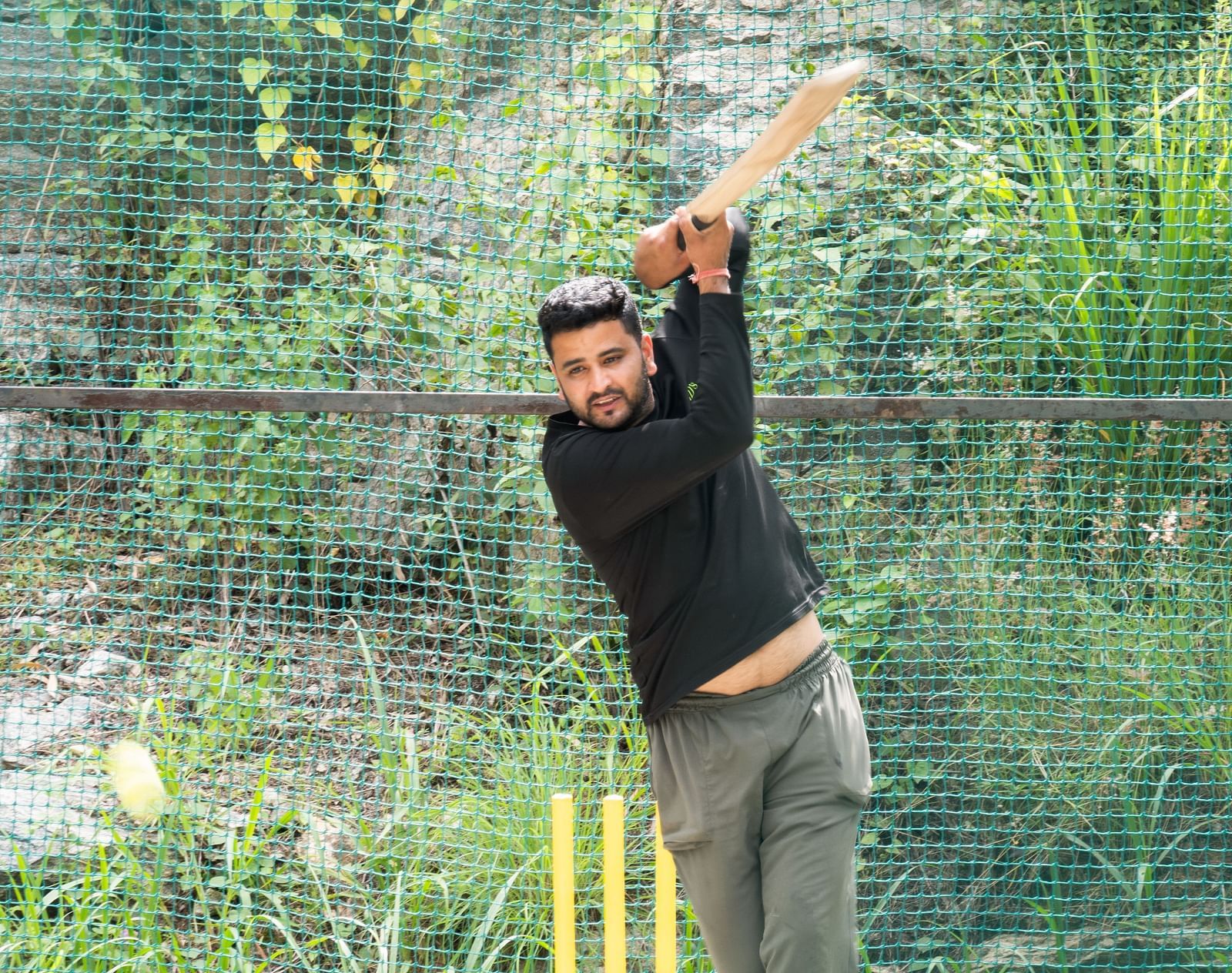 A person swings a cricket bat in a net practice area, surrounded by green fencing | RD's Nature Retreat, Bangalore