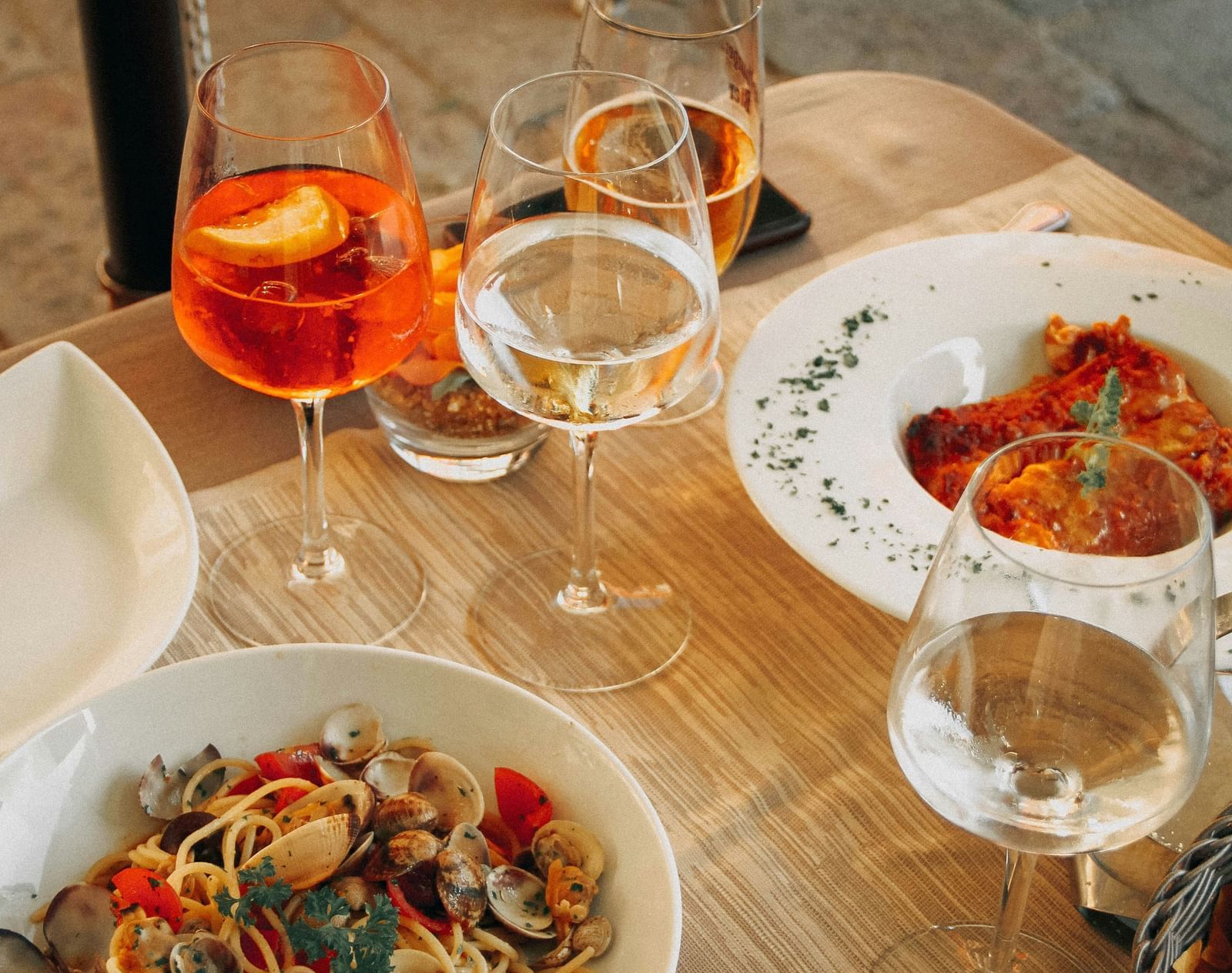 A close-up, slightly blurred shot of a dinner setting on a wooden table, showing a bowl of pasta or salad, a glass of orange-coloured drink, and a glass of water.