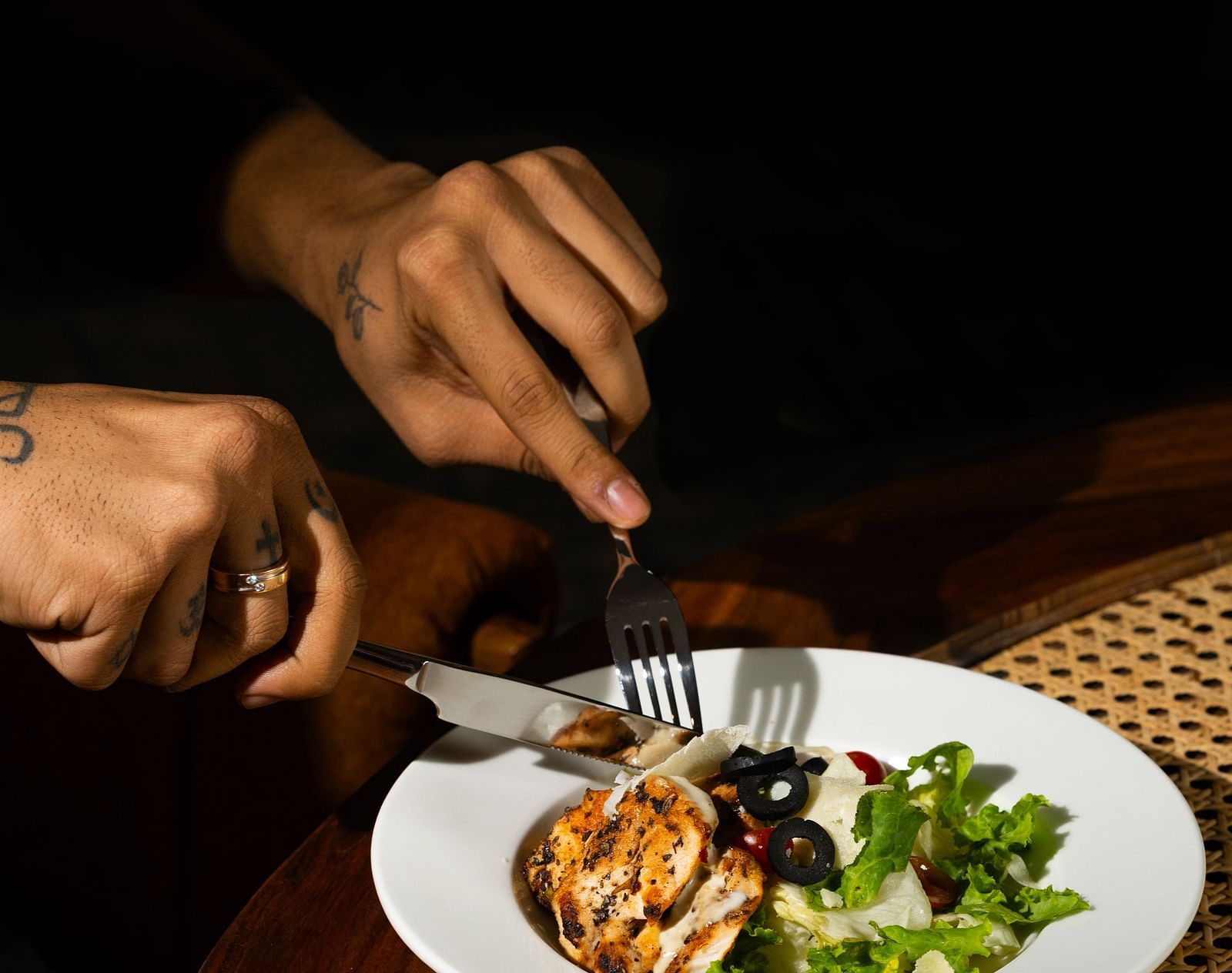A person's hands holding utensils over a plate of food, set on a table with a woven placemat at The Qualia Resort Club & Brewery, Udaipur.