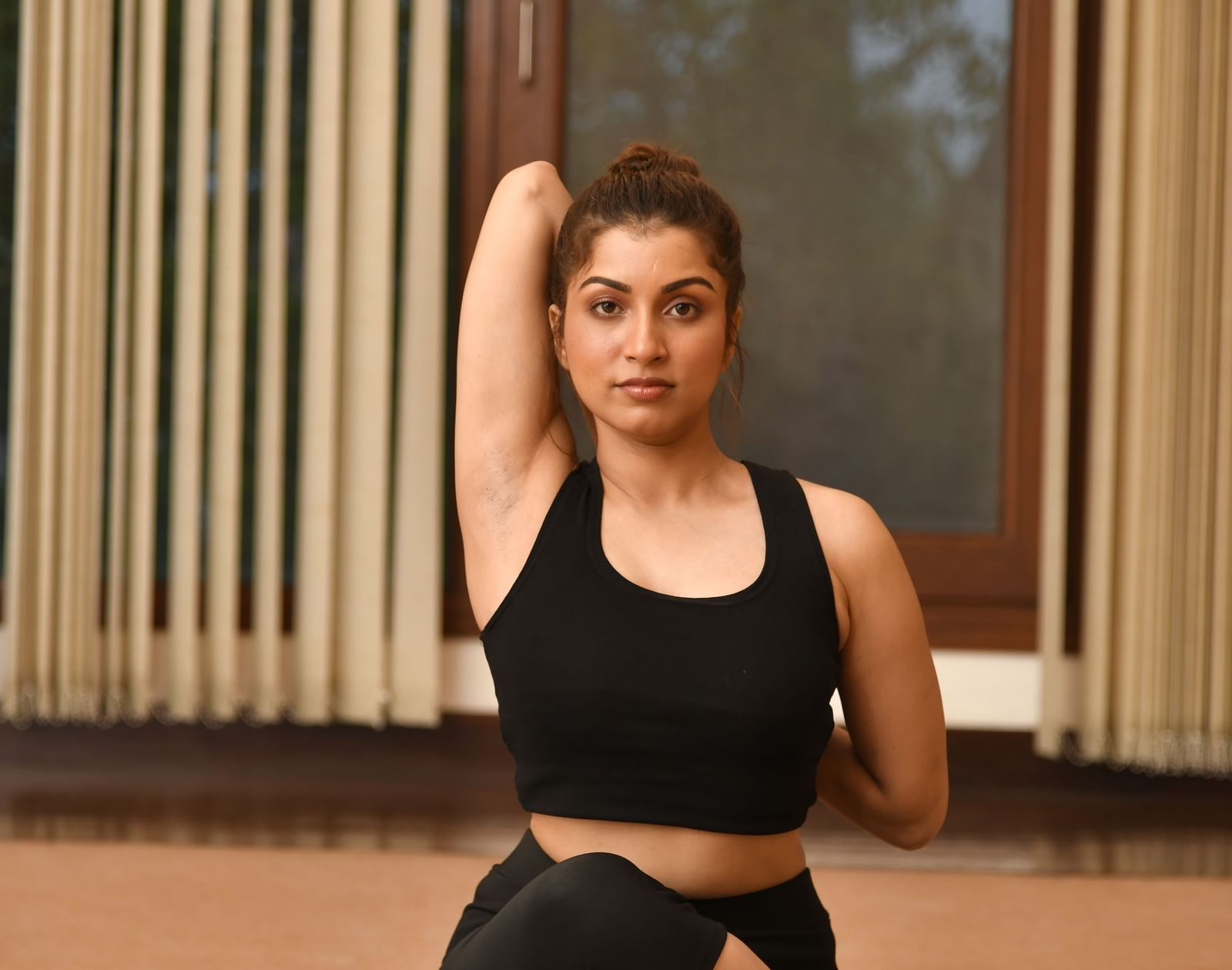 A woman in black activewear performs a seated yoga pose, twisting her torso and looking towards the camera, in a room with large windows