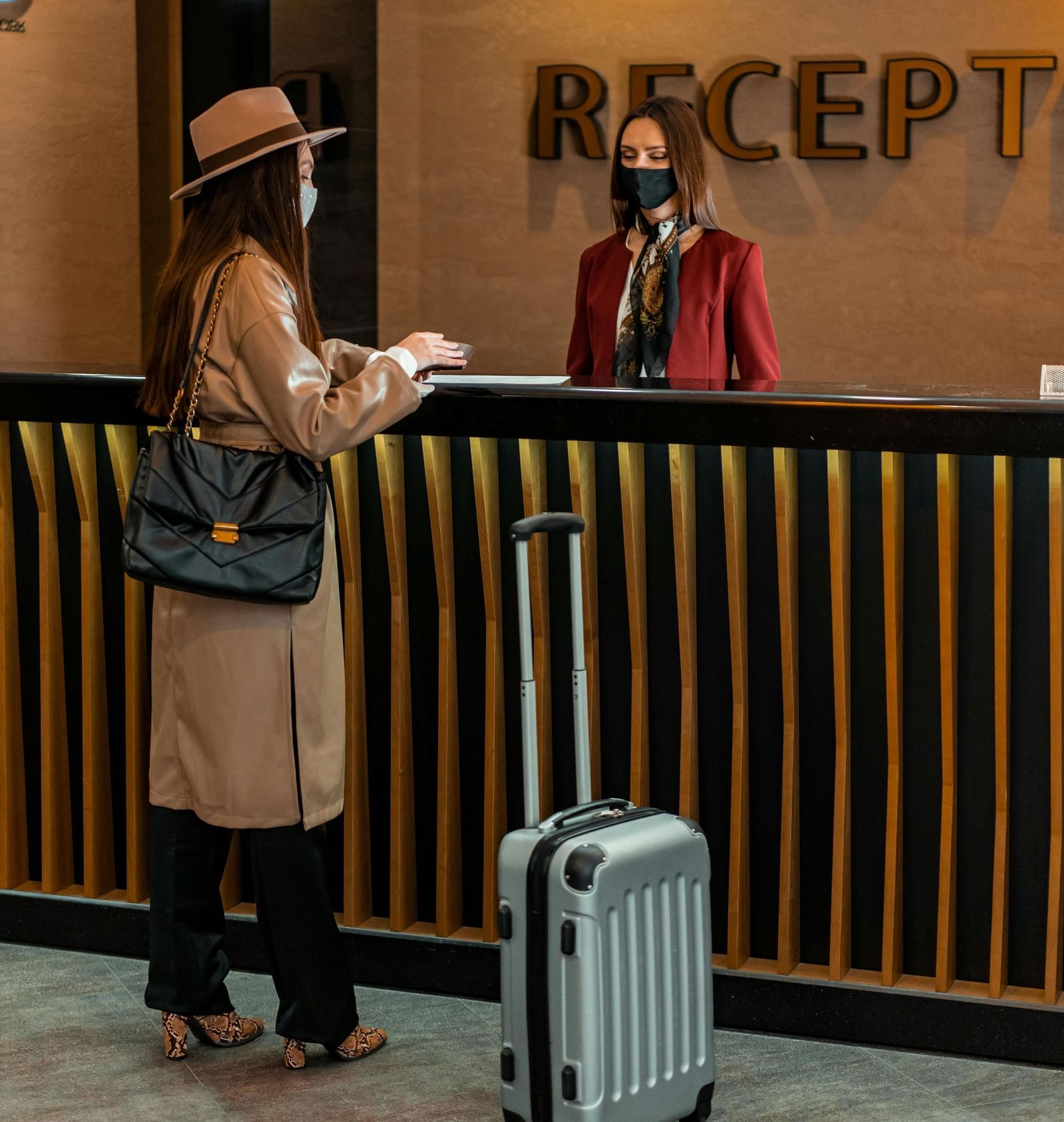 A receptionist standing behind the desk greeting the guest carrying a luggage trolley in front of her.