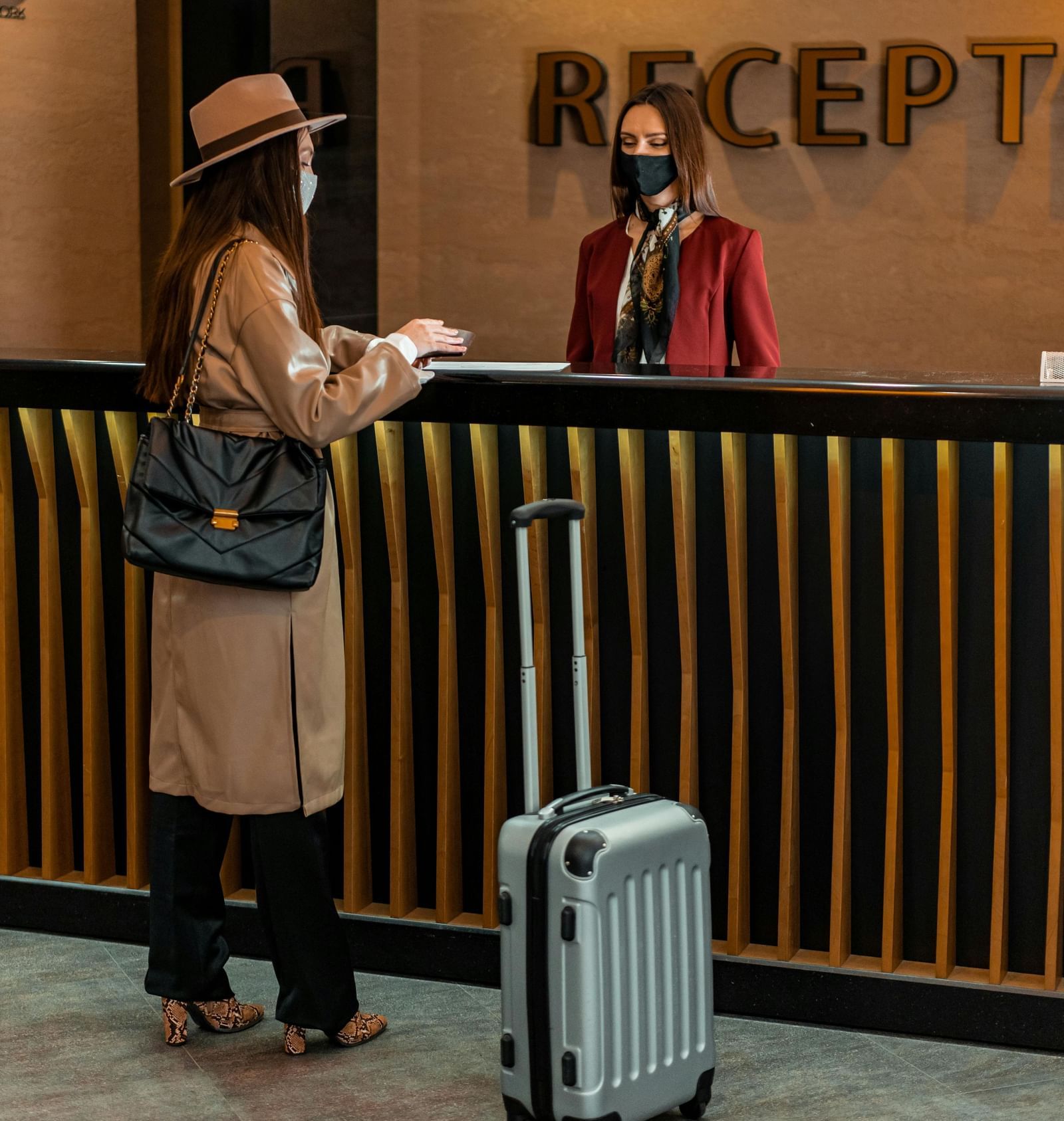 A receptionist standing behind the desk greeting the guest carrying a luggage trolley in front of her.