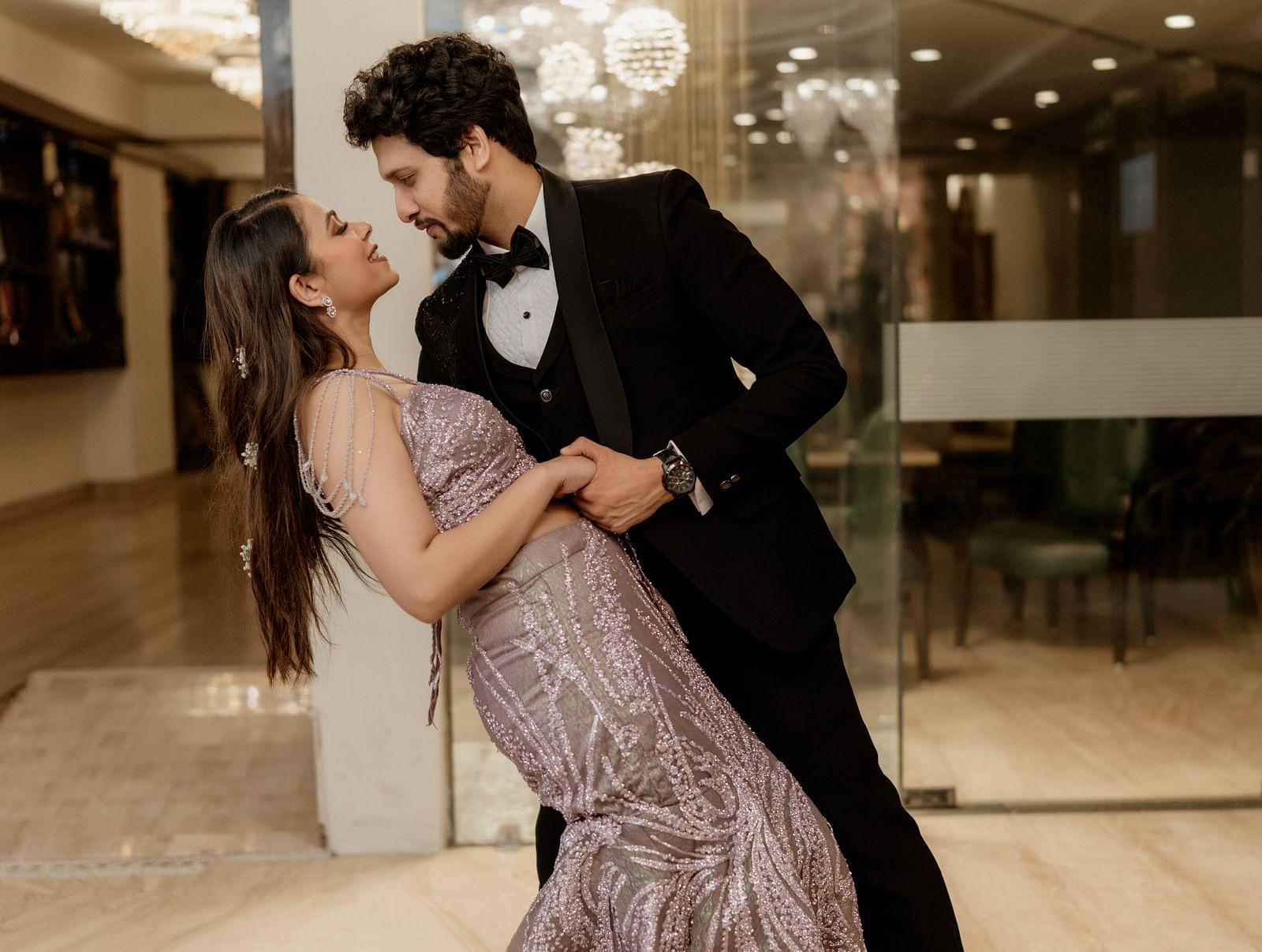  couple is captured in a dance pose, the man in a dark suit and the woman in a flowing gown, set against a bright, modern interior.