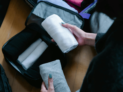 Hands packing neatly folded clothes into a suitcase on a wooden floor.