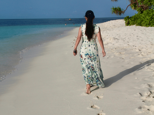 A person walking along a sandy beach near the shoreline, with palm trees on one side and ocean water in the background