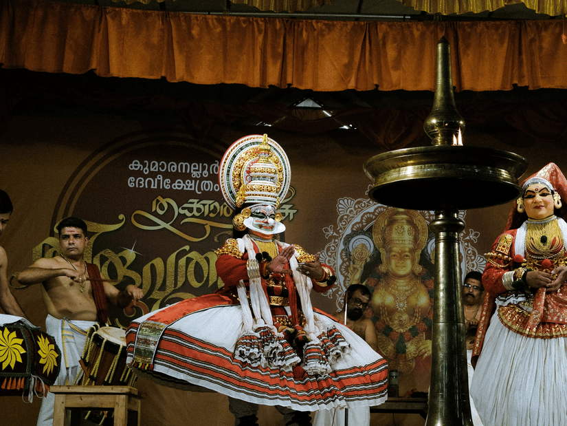 Kathakali dancers in vibrant, ornate attire perform on stage alongside percussionists and a large brass oil lamp.