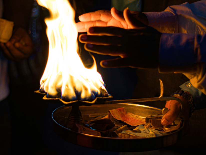Hands cupped above a bright, flaming brass diya lamp during a dark ceremonial evening ritual or aarti at Kali Ghat Kali Mandir