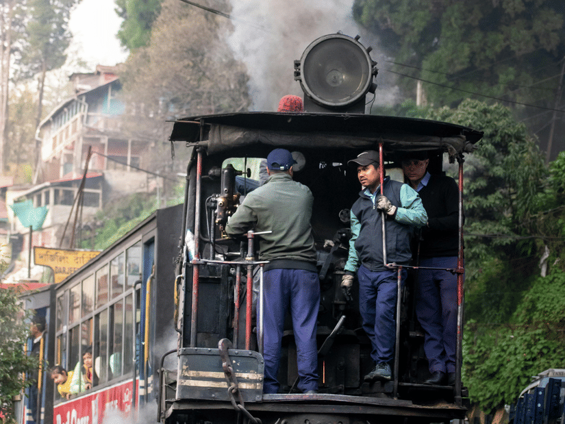 The iconic Darjeeling Himalayan Railway steam engine with billowing smoke, passing through lush hills and narrow tracks.