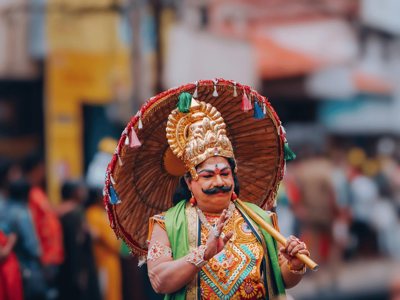 Performer wearing an ornate traditional costume and painted mask participates in a lively Kerala cultural procession through a busy street