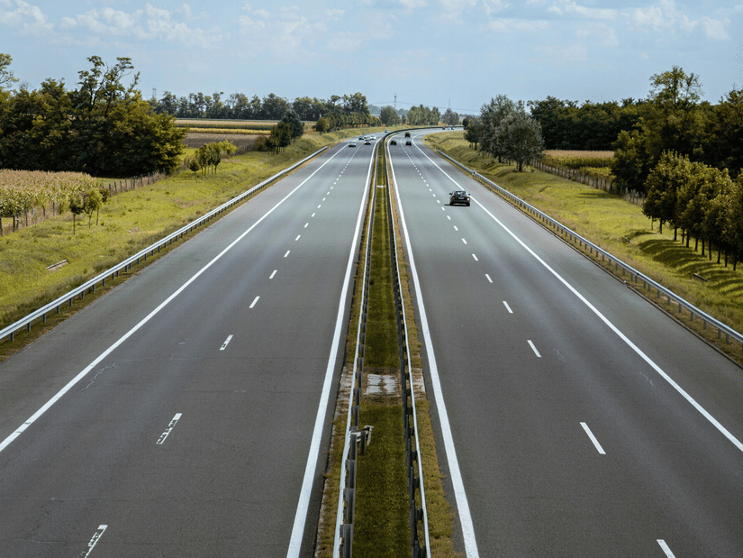 A view atop of a four lane highway divided by a small lawn, cars travelling on the road and scattered trees on either side of the road.