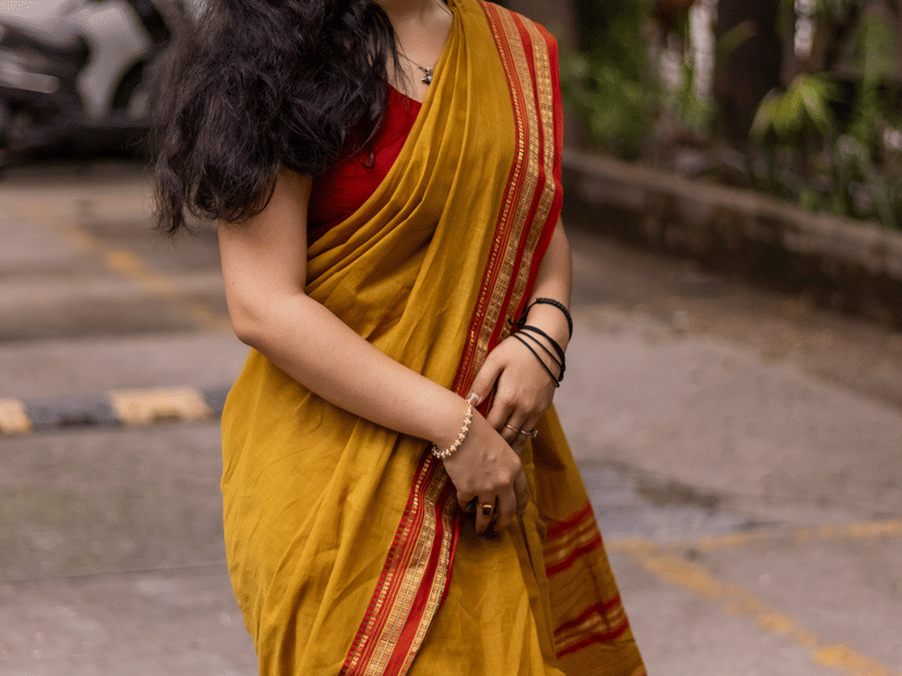 A woman wearing a mustard-yellow cotton saree with an orange border smiles while standing outdoors on a peaceful tree-lined pathway.