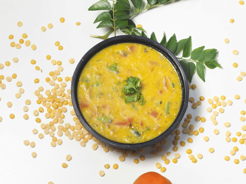 A top down view of a bowl of Dal Kichdi kept next to curry leaves, a tomato, and lentils sprayed around it.