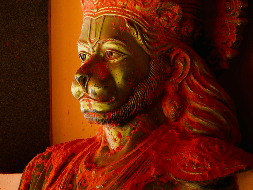 A red-orange statue of a Hindu deity with an ornate crown and detailed facial features, lit against a dark background.
