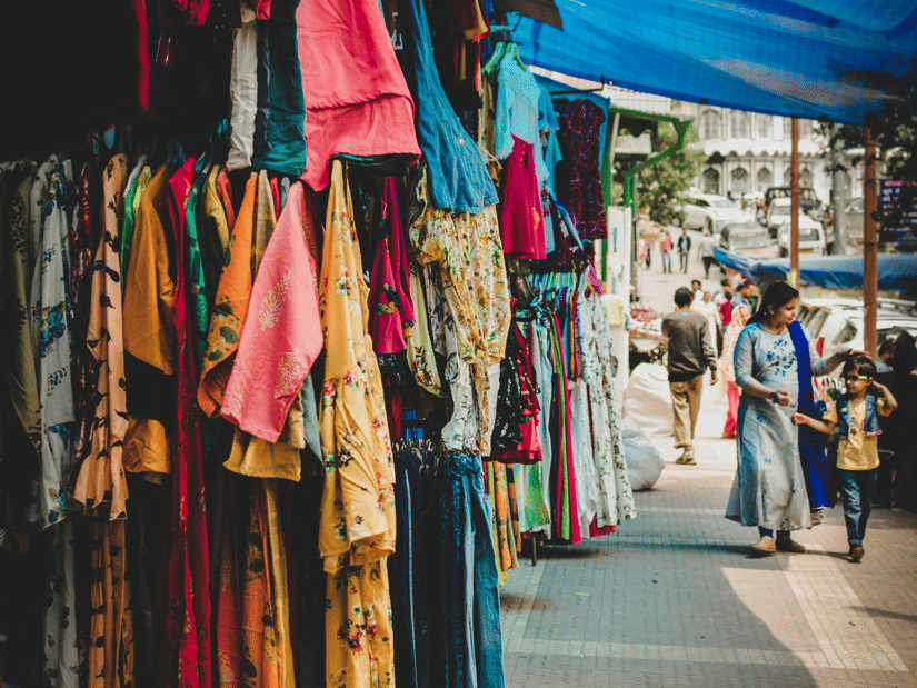 Colorful clothing hanging on display at an outdoor market stall under a blue tarp, with people walking on a sidewalk nearby.