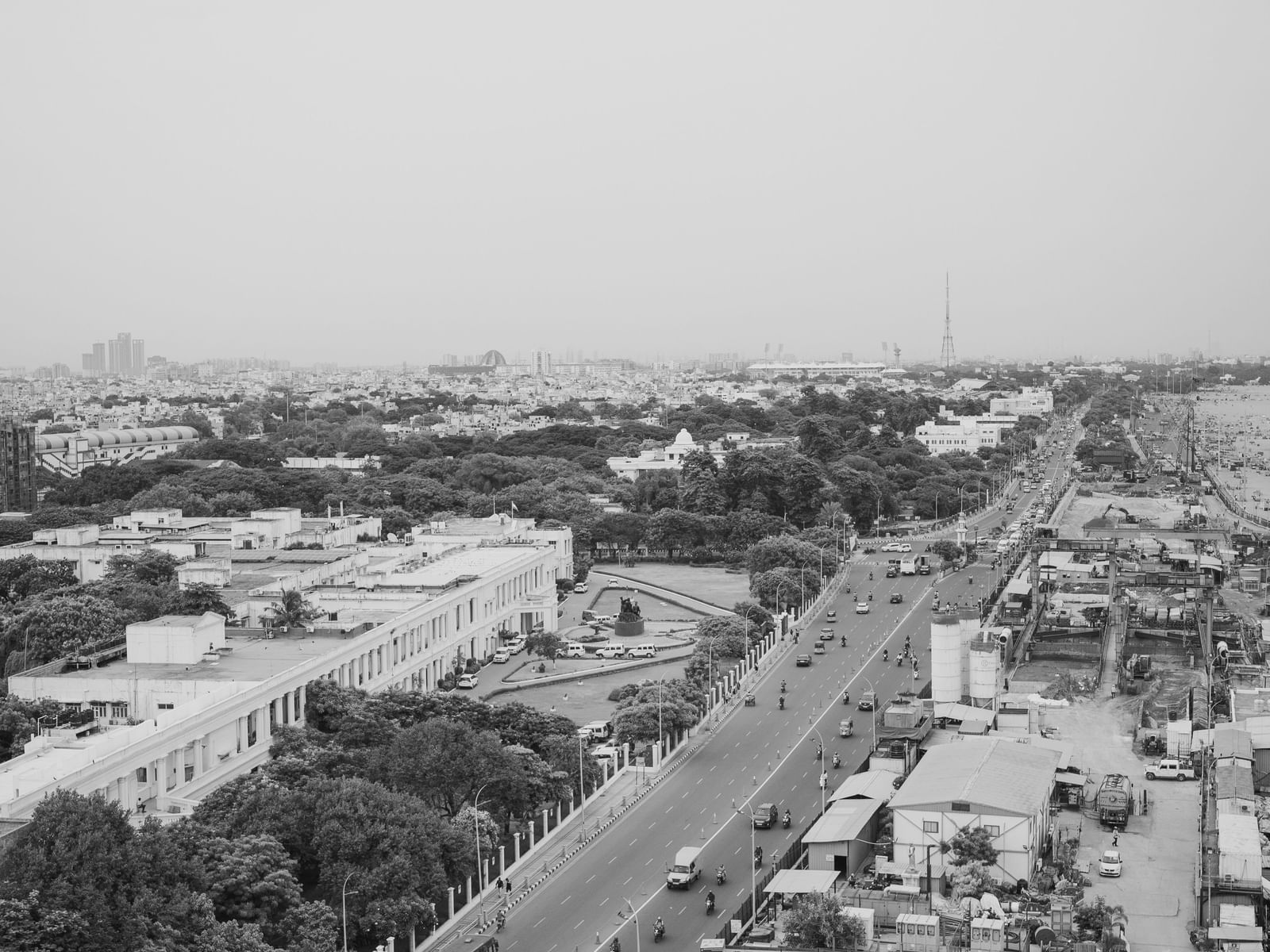 A black and white aerial photograph of Chennai shows a busy main road running through the city, flanked by residential and commercial buildings, trees, and dense urban infrastructure, with vehicles visible and the skyline extending into the distance under 