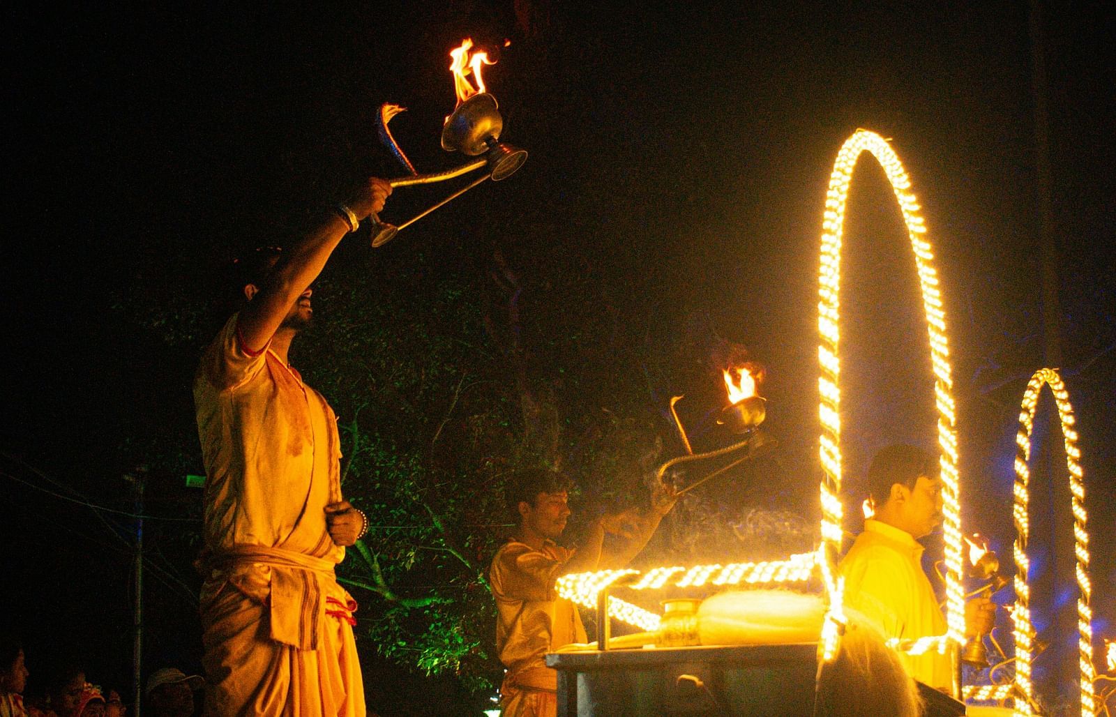 three priests doing aarti during late evening as the devotees are sitting in the background