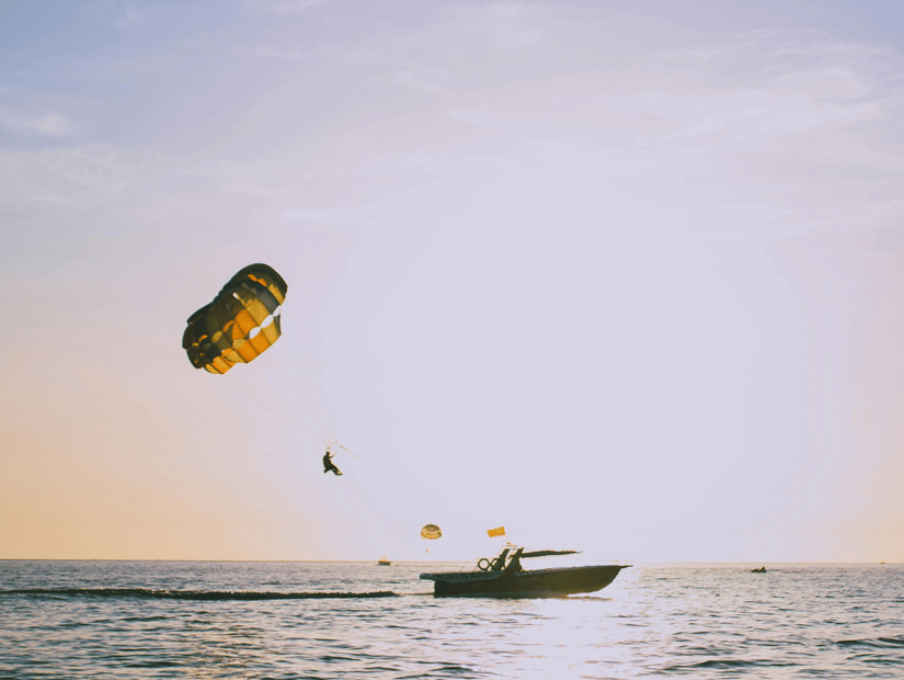A person parasailing with a colorful parachute over calm sea waters during golden hour with a motorboat visible on the water surface and a soft pastel sky in the background.