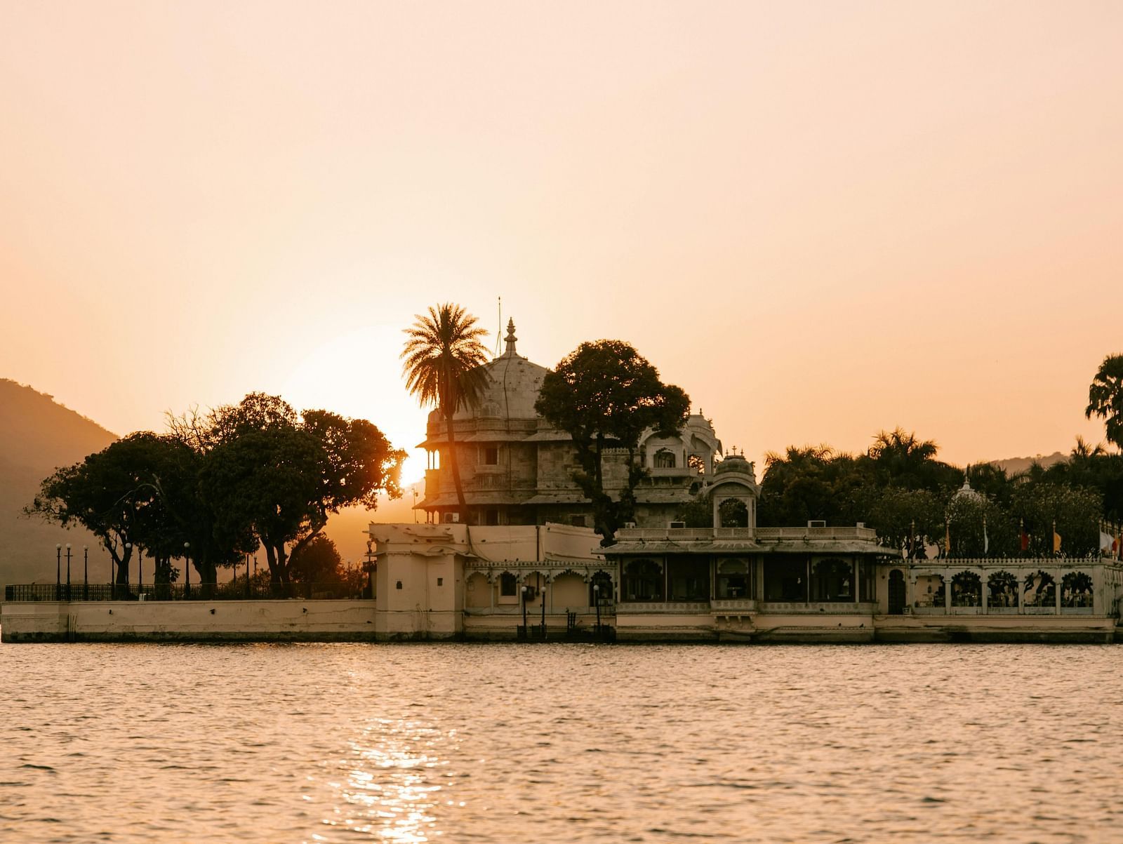 White building with royal architecture surrounded by trees on a water body at sunset.
