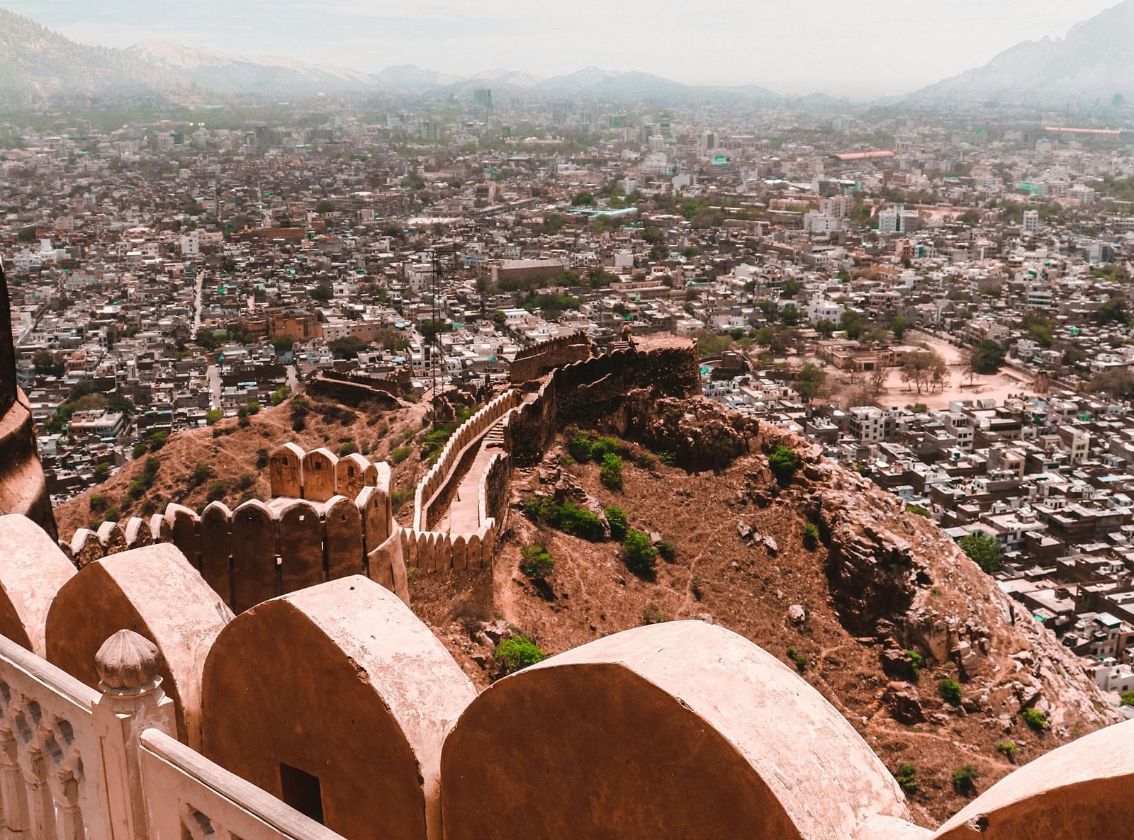 A high-angle view from Nahargarh Fort, Jaipur, overlooking the sprawling city with its densely packed buildings and a distant mountain range under a cloudy sky.