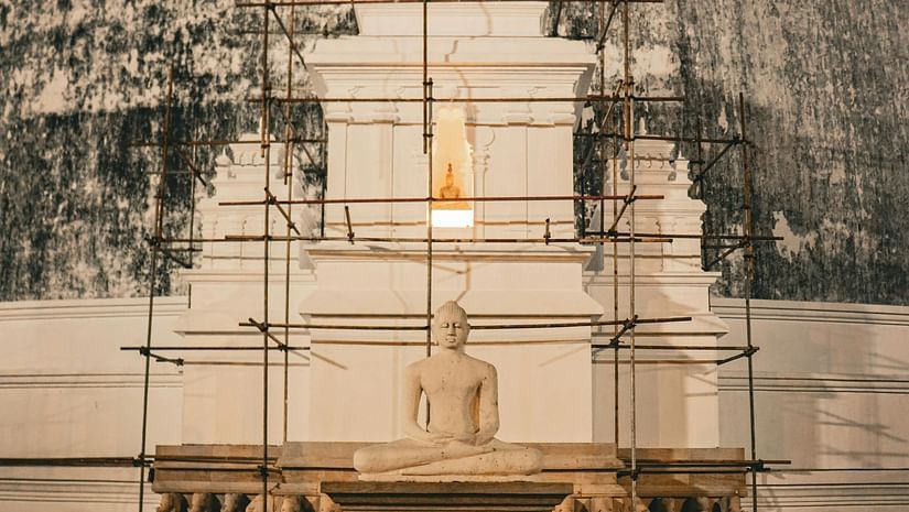 A low-angle shot of a large, white Buddhist stupa with scaffolding against a dark background, featuring people praying at the base.