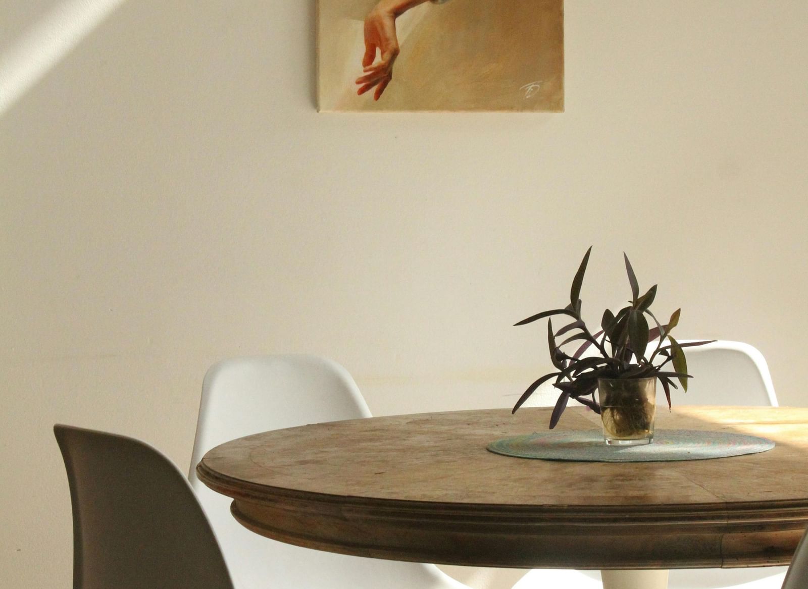 A simple dining area features a round wooden table with chairs, positioned near a white wall with a framed picture.