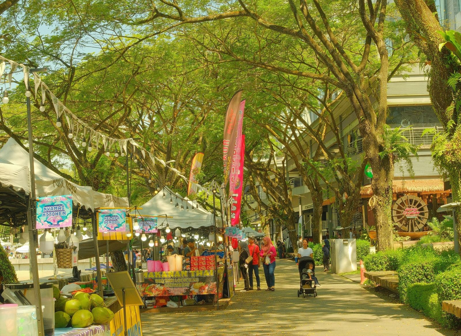 A bright, sunlit pedestrian pathway lined with trees and small market stalls or shops.