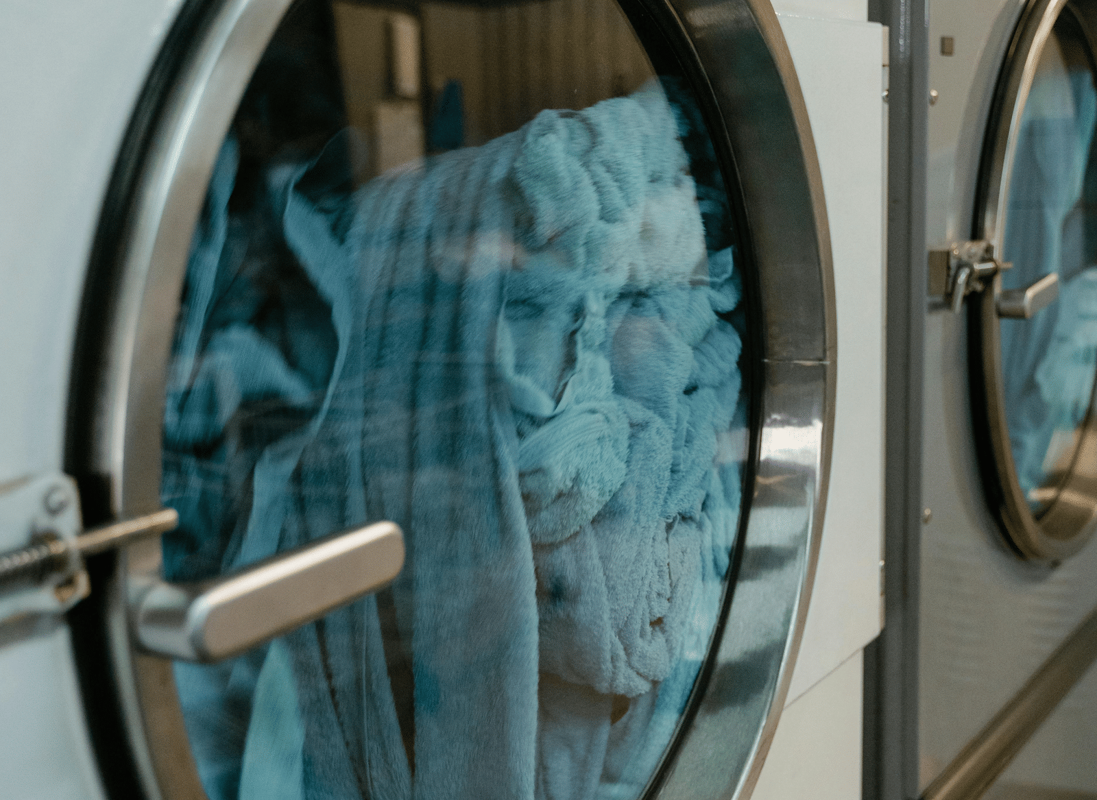 A close-up view of a metal commercial washing machine door with a round glass window, showing laundry rotating inside.