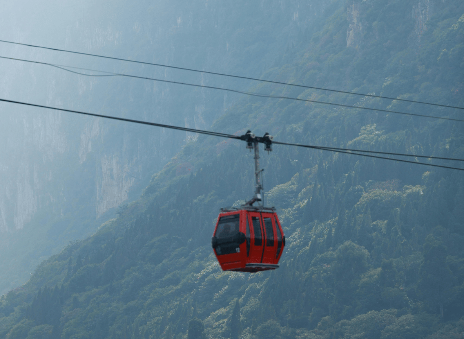 A red cable car gondola suspended on a cable, travelling high above a green, misty valley