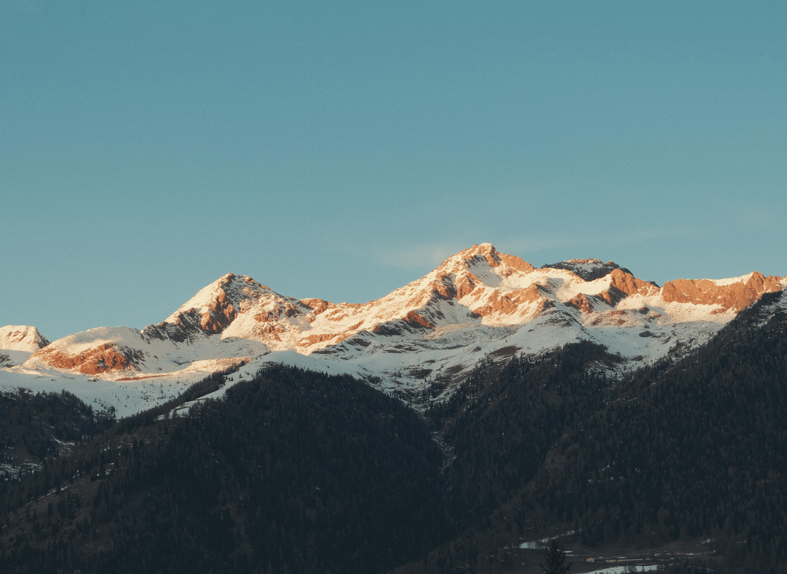 A mountain range with snow-capped peaks visible above a dark foreground, set against a clear blue sky
