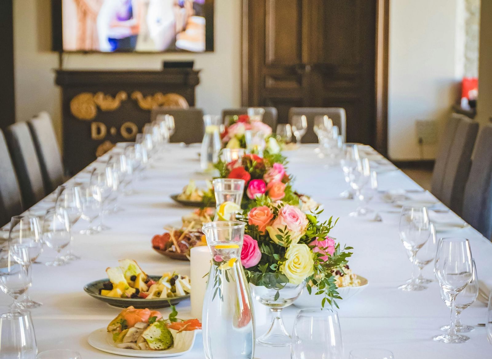 A long dining table decorated with flowers in the middle along with shiny glassware and cutlery set on the table.