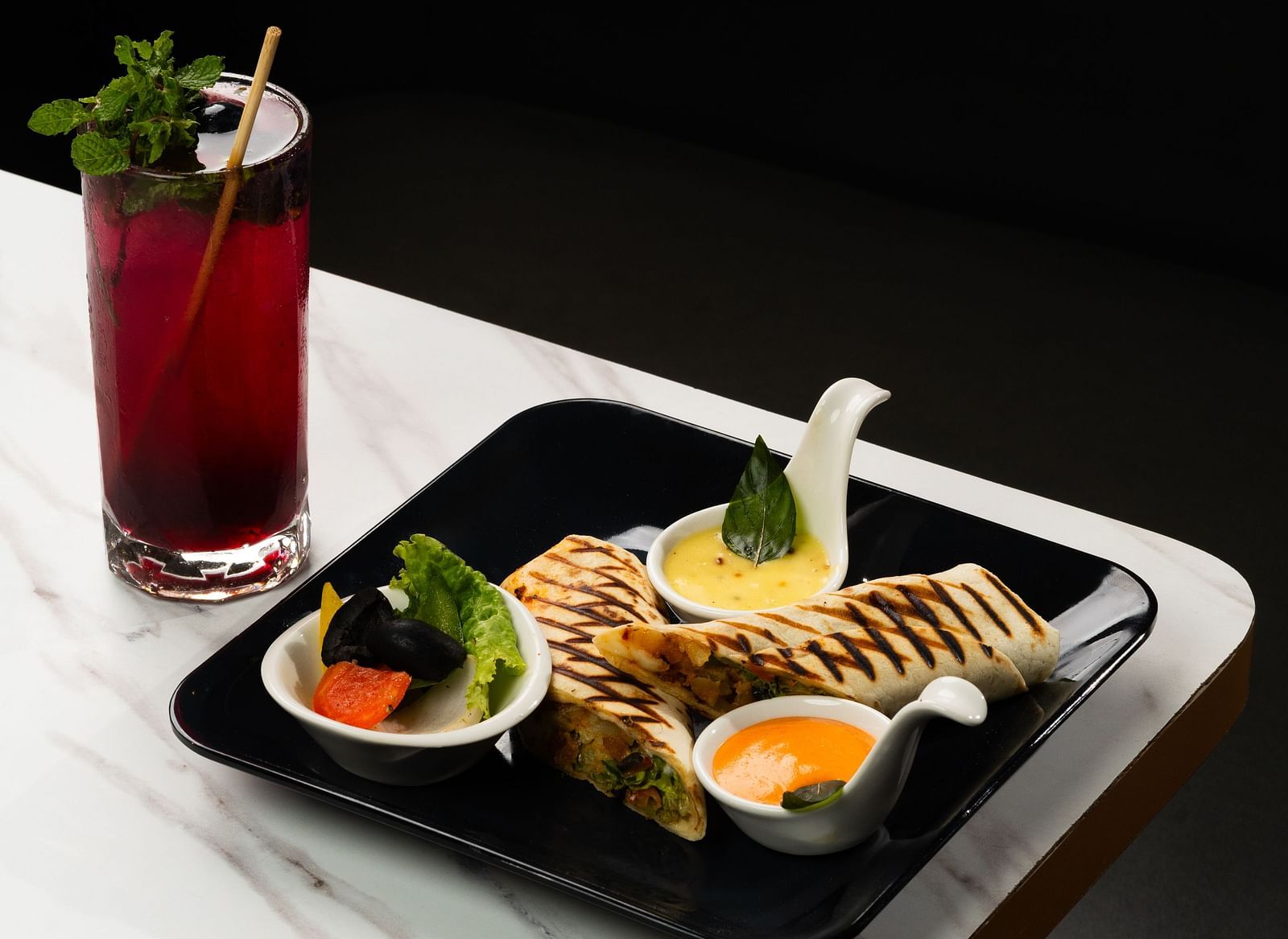A dark and moody food presentation with a red drink and a tray of appetizers on a white tablecloth at The Qualia Resort Club & Brewery, Udaipur.