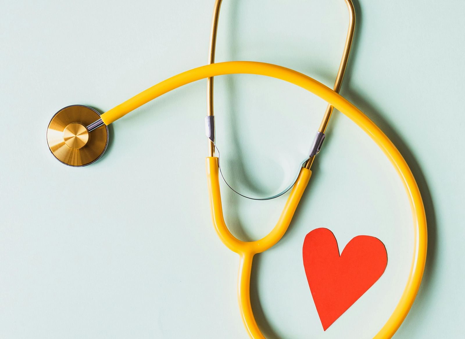 A yellow stethoscope forming a heart shape with a red paper heart cutout inside, against a light blue background.
