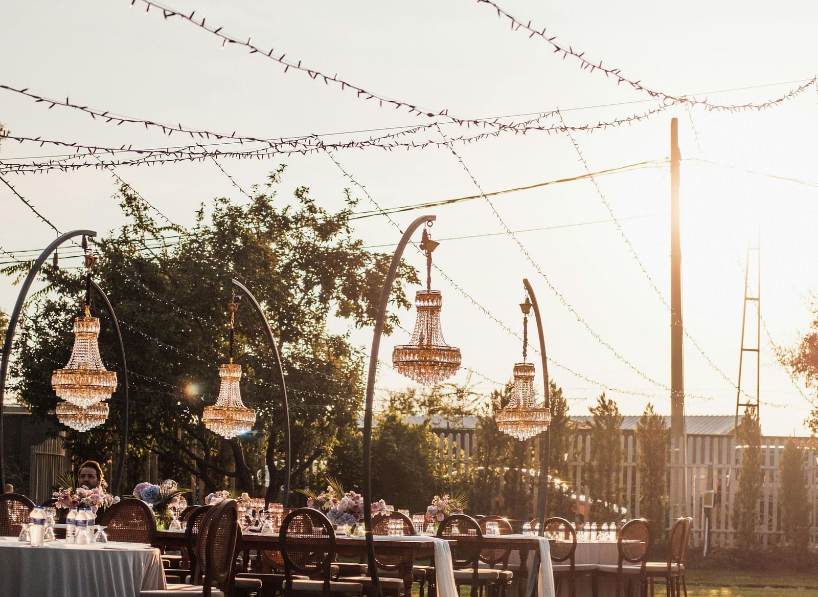 Outdoor event setup with long tables, chandeliers hanging from trees, and string lights overhead.
