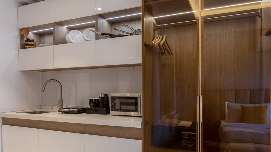 Vertical view of a compact kitchen area adjacent to a tall, tinted glass wardrobe in a luxury room.