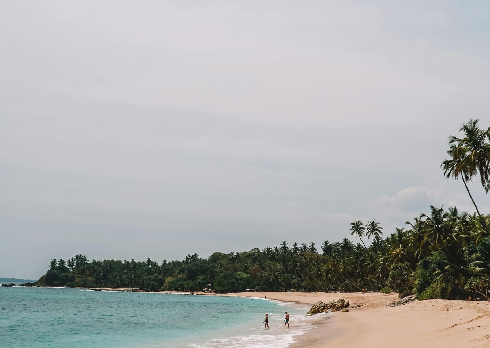 Quiet tropical beach with gentle waves, distant figures walking along shore, and palm trees lining the sandy coastline.