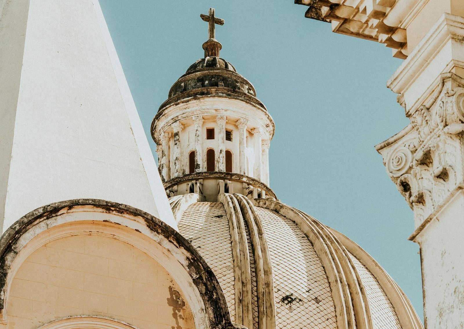 A close-up shot of the weathered dome and cross of a light-colored cathedral, set against a pale blue sky.