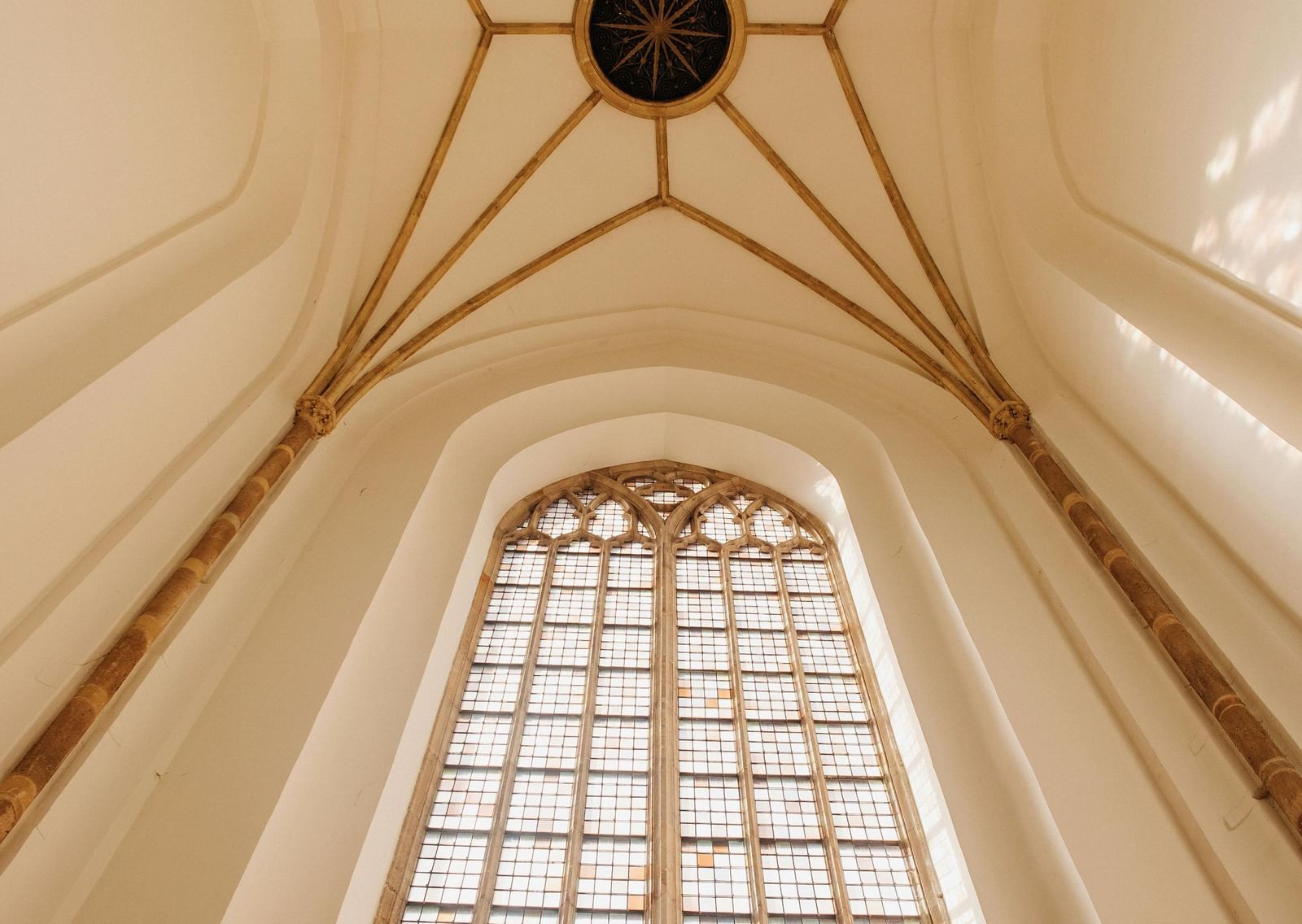 A low-angle shot of a cathedral's vaulted ceiling and large stained-glass window. 
