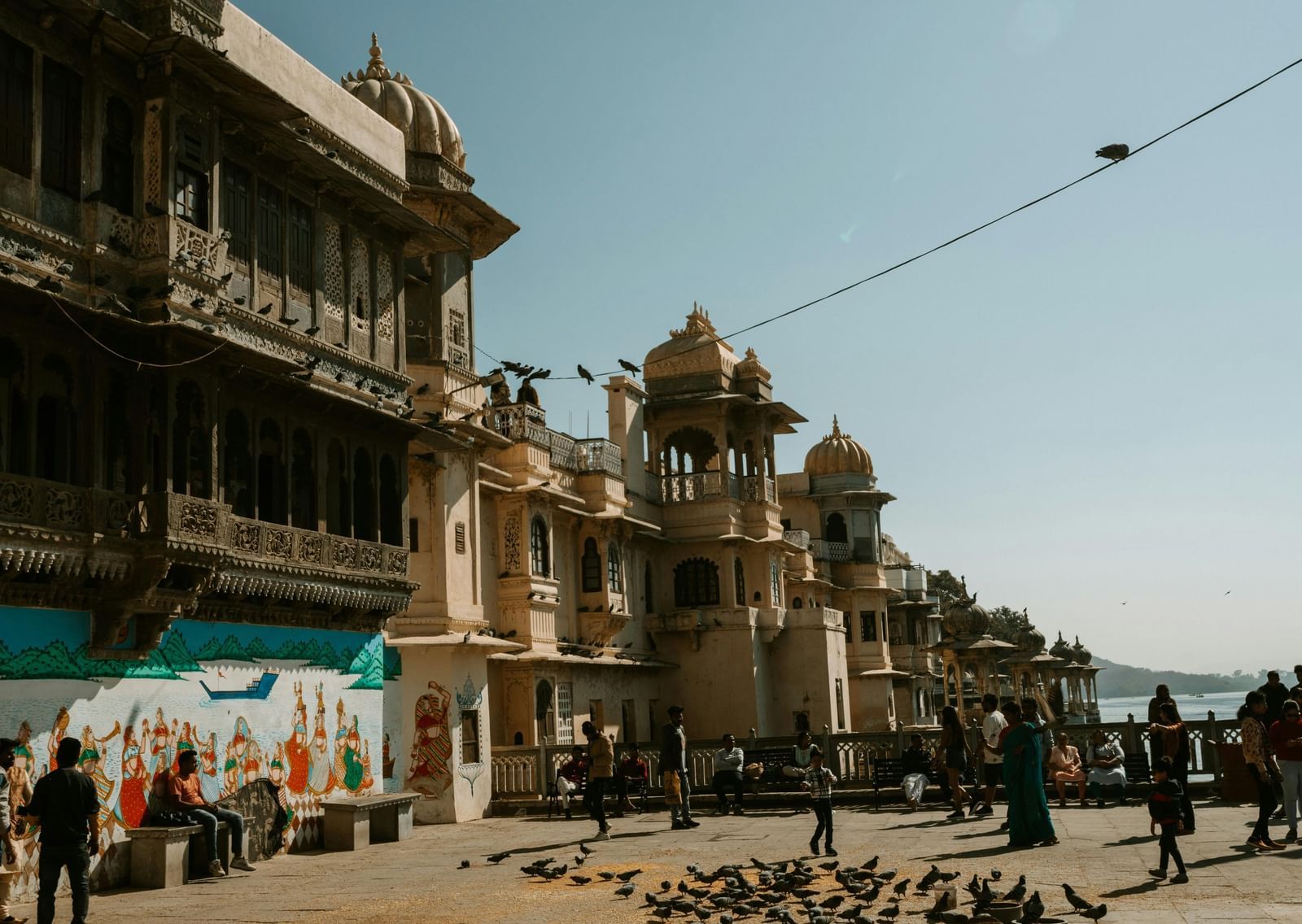 Streets of Rajasthan with people walking around and a flock of birds on ground under a blue sky.