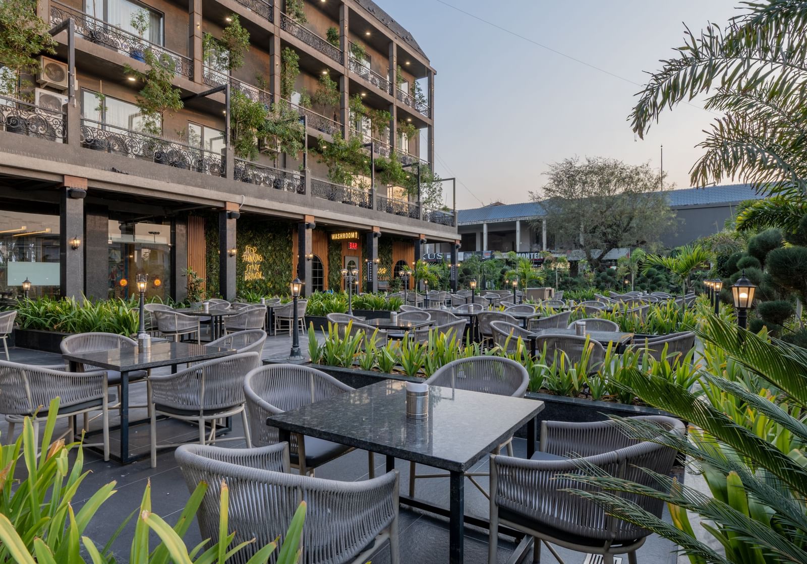 Outdoor seating area at Hotel Highway King, Bagru, with tables and chairs surrounded by plants and hotel balconies.