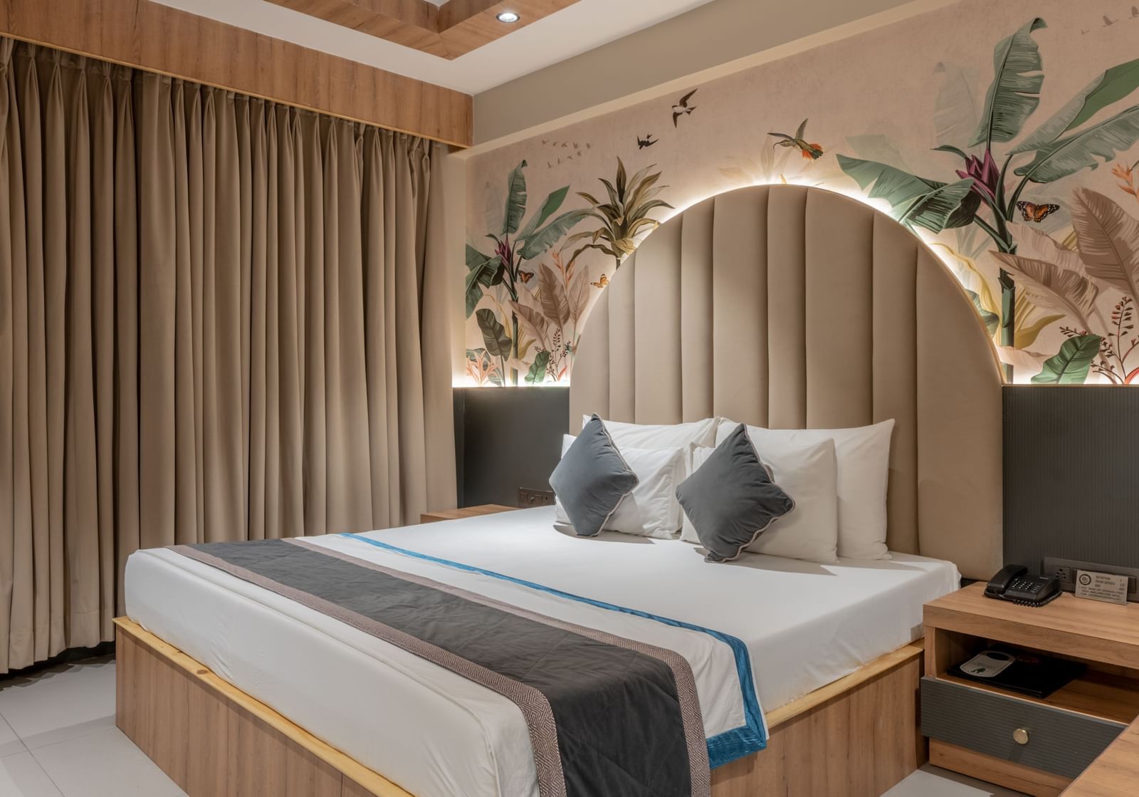 A bedroom with king size bed, ceiling fan, a wooden side table, and curtain spread across the window at Hotel Highway King, Jaipur.