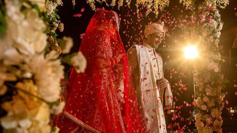 A bride in a red lehenga stands under a beautifully decorated floral canopy, her face covered with a traditional veil, surrounded by lush flowers and soft lighting.