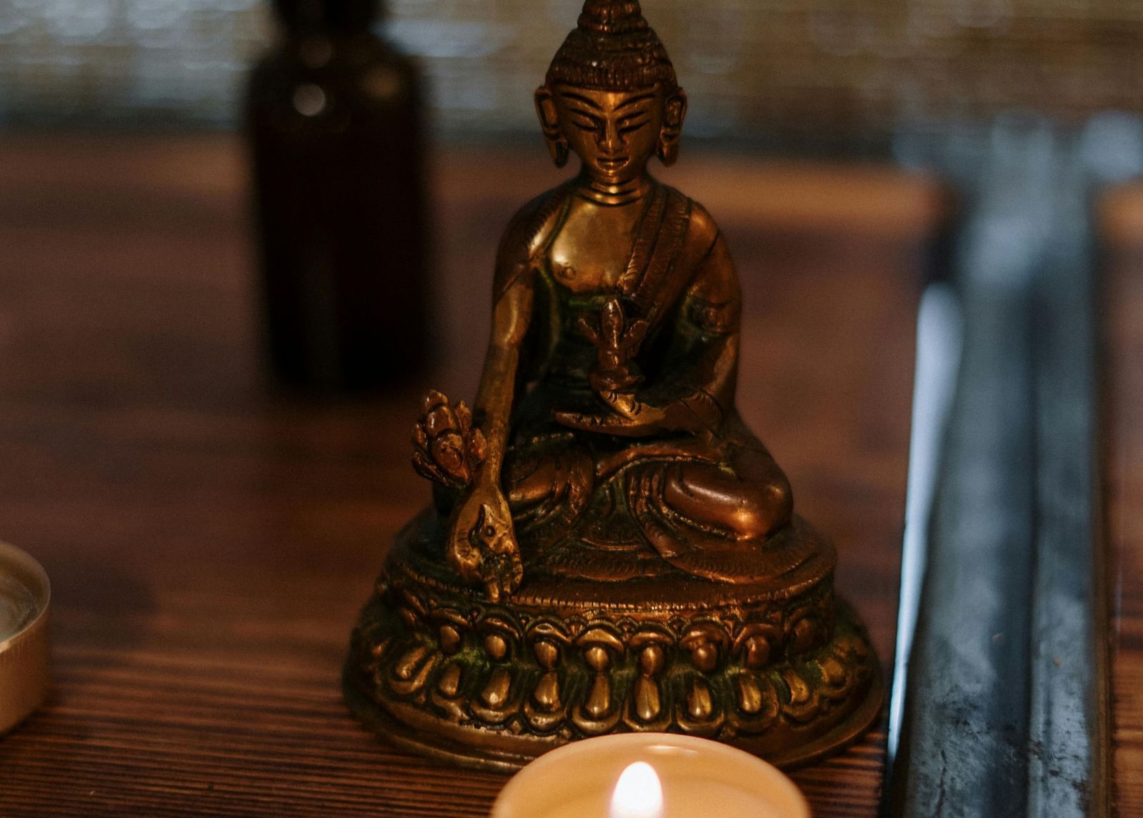 Small golden Buddha statue, two lit tealight candles, and a dark glass dropper bottle on a wooden surface.