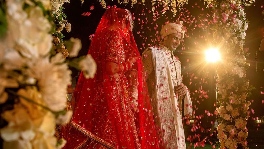 A bride in a red lehenga stands under a beautifully decorated floral canopy, her face covered with a traditional veil, surrounded by lush flowers and soft lighting.
