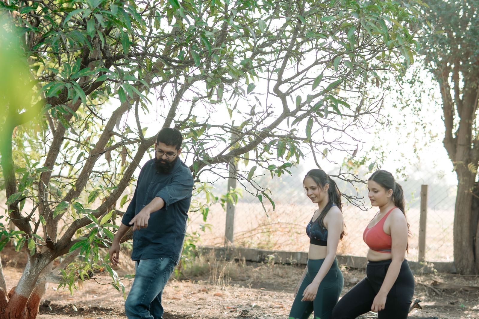 A man pointing towards plants and two women looking at the same spot amidst the farm covered in plants and trees at Daksh Eden Greenz in Sasan Gir.