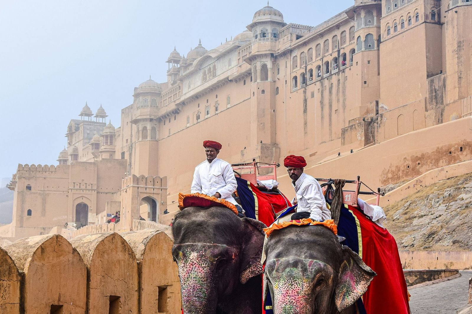 Two elephants adorned with colourfull cloths carry riders up a path towards a large fort with stone walls.