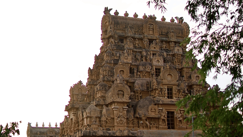 A large, ancient stone temple with multiple tiers and intricate carvings is partially visible through green foliage.