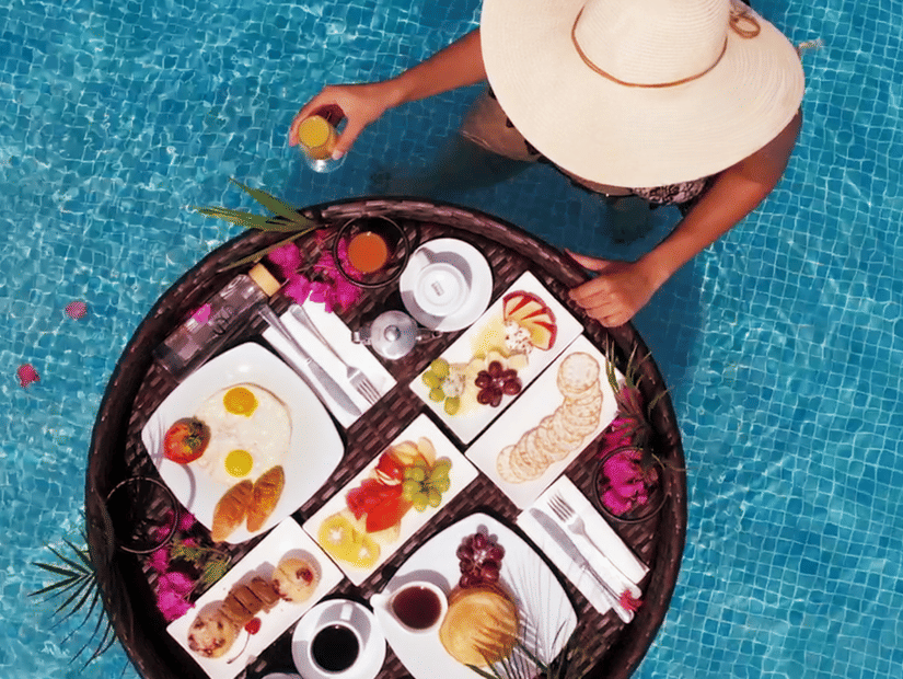 A person wearing a wide-brimmed hat enjoying a floating breakfast tray with assorted dishes and drinks in a swimming pool