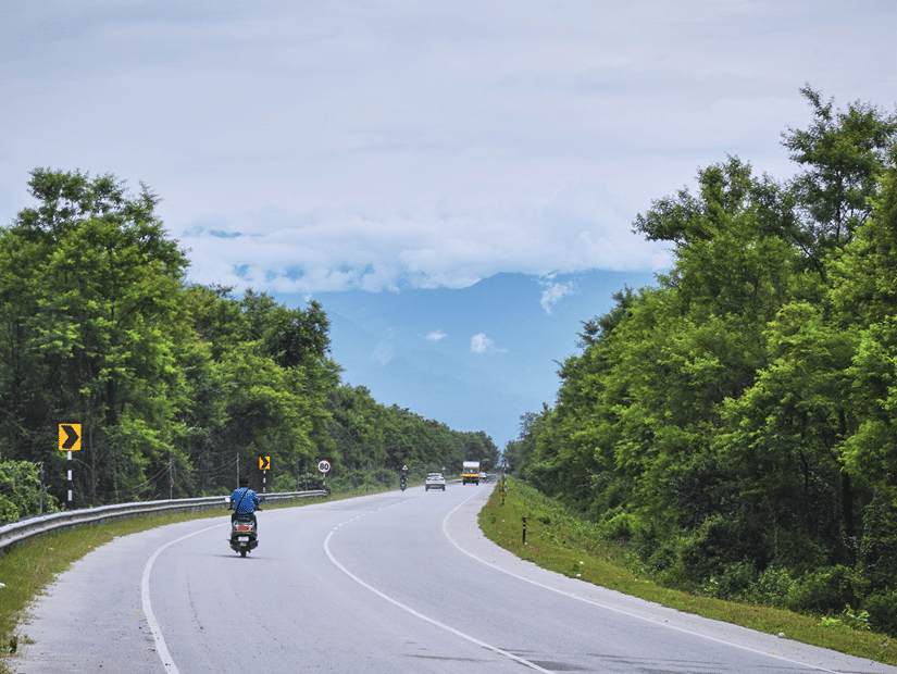 A highway curve with greenery lining up its 2 sides and a man driving a scooter down the road under a clear blue sky.